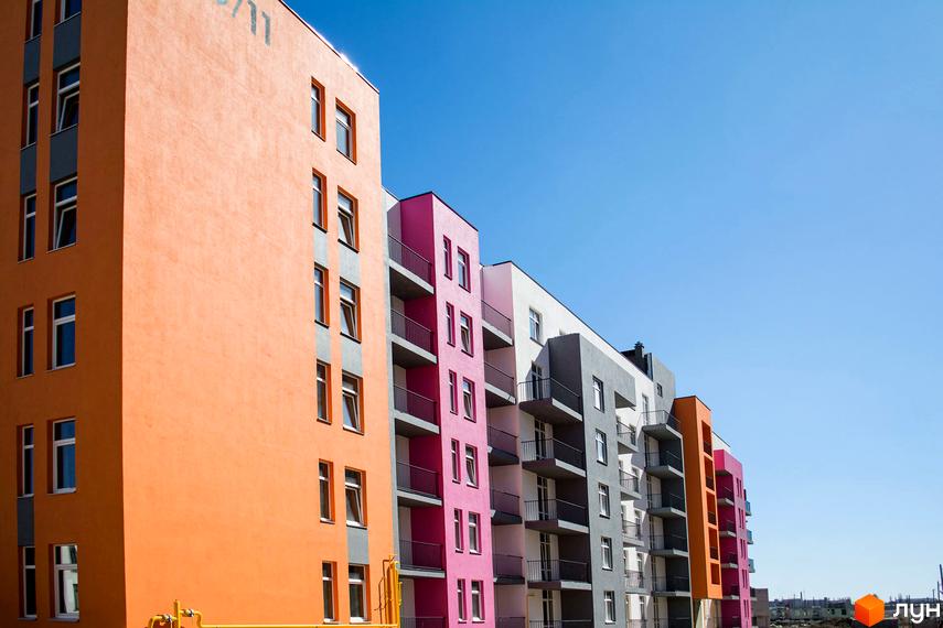 Фасад житлового комплексу Європейка з різнокольоровими секціями: помаранчевою, рожевою, білою та сірою. Будівлі мають багато вікон та балконів, розташовані під яскравим блакитним небом.