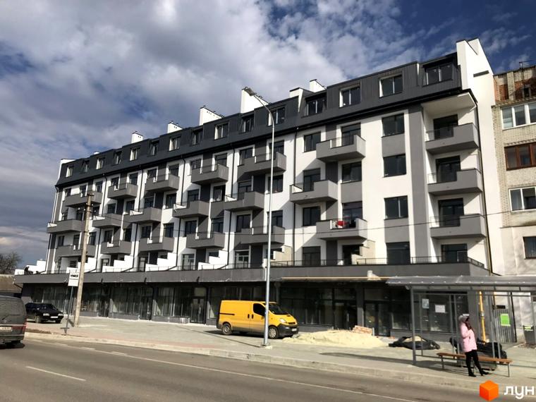 Новобудова ЖК Європа з сірими та білими фасадами, балконами та комерційними приміщеннями на першому поверсі. Поруч дорога, зупинка громадського транспорту та припарковані автомобілі.