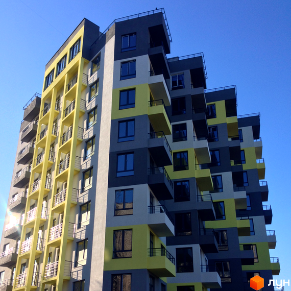 Житловий комплекс Енергія плюс. Фасад новобудови з жовтими та сірими елементами, балконами та панорамними вікнами на тлі блакитного неба.