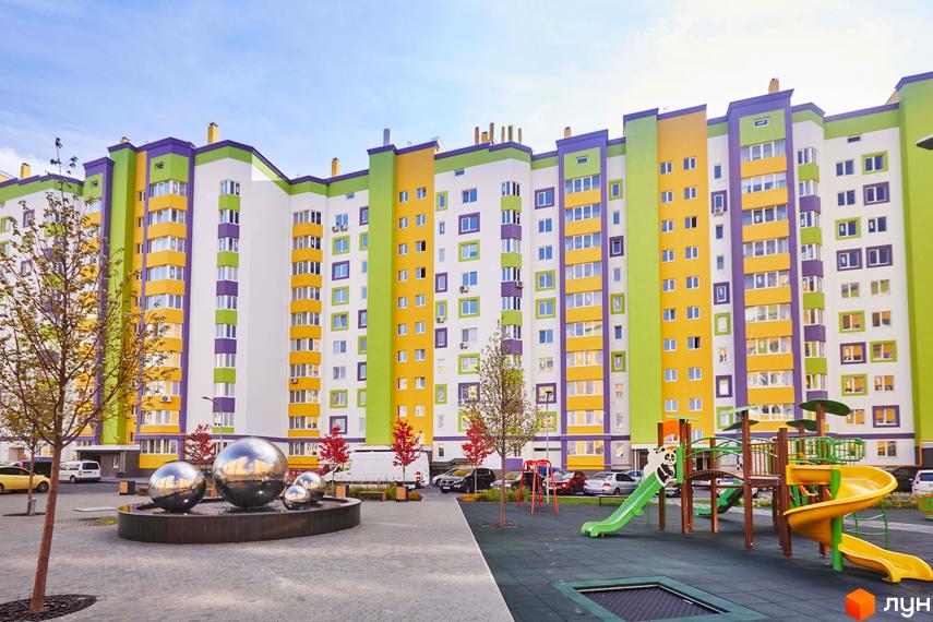 Житловий комплекс Щасливий на Яблуневій. Багатоповерховий будинок з яскравим фасадом у жовтих, зелених та фіолетових кольорах. Поруч дитячий майданчик та зона відпочинку з фонтаном.