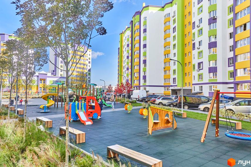 Житловий комплекс Щасливий на Яблуневій: яскраві багатоповерхові будинки з кольоровими фасадами. Дитячий майданчик з гірками, гойдалками та пісочницею, зелена зона з деревами та лавками.