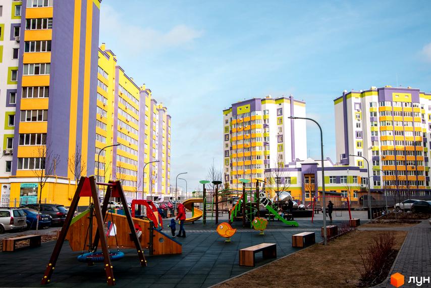 Житловий комплекс Щасливий на Яблуневій: яскраві багатоповерхівки з фіолетовими та жовтими фасадами. Сучасний дитячий майданчик з гірками та гойдалками перед будинками.