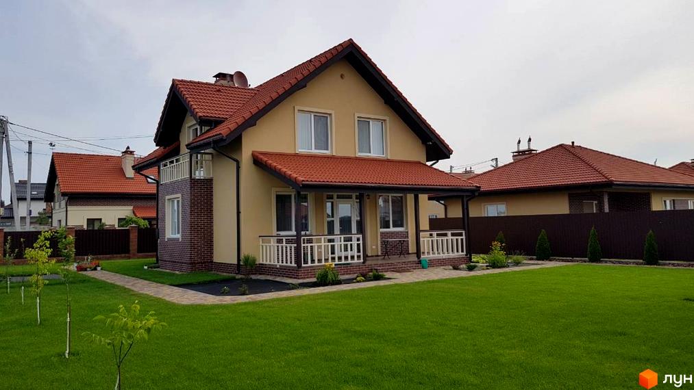Котеджне містечко Сонячний: двоповерховий будинок світло-жовтого кольору з червоним черепичним дахом, балконом та ганком. Навколо будинку доглянутий зелений газон та доріжка з бруківки.