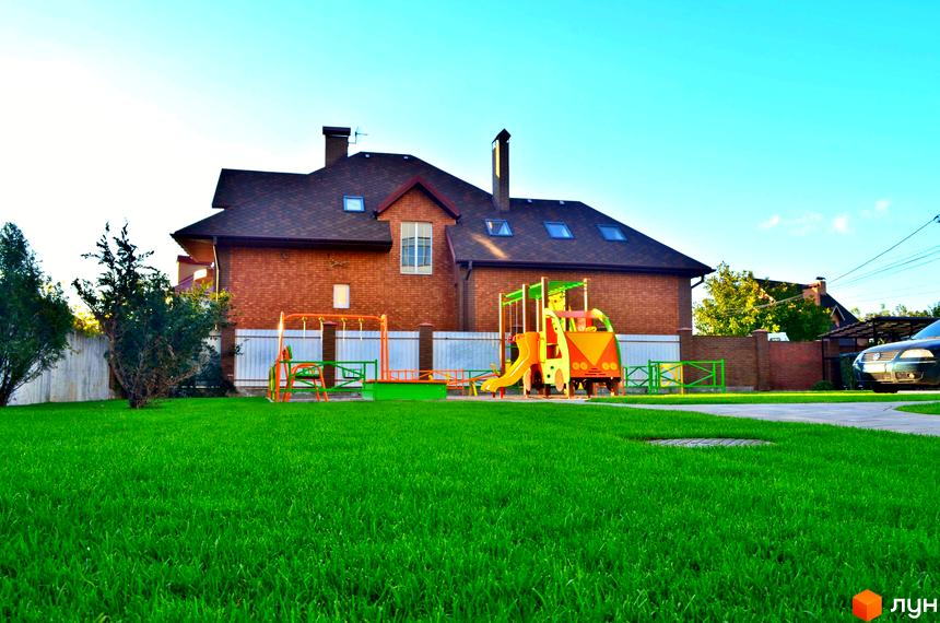 Цегляний будинок з темно-коричневим дахом та дитячим майданчиком на прибудинковій території. На газоні розташовані гойдалки та гірка.