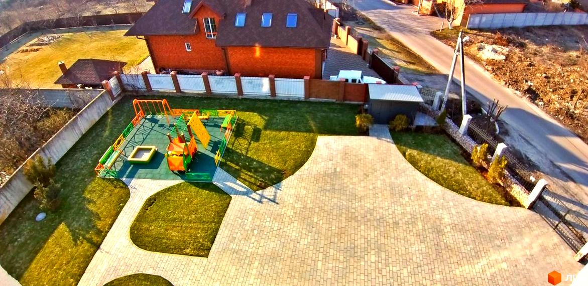 Цегляний будинок з коричневою черепицею та мансардними вікнами. На прибудинковій території розташований дитячий майданчик та вимощена бруківкою доріжка.