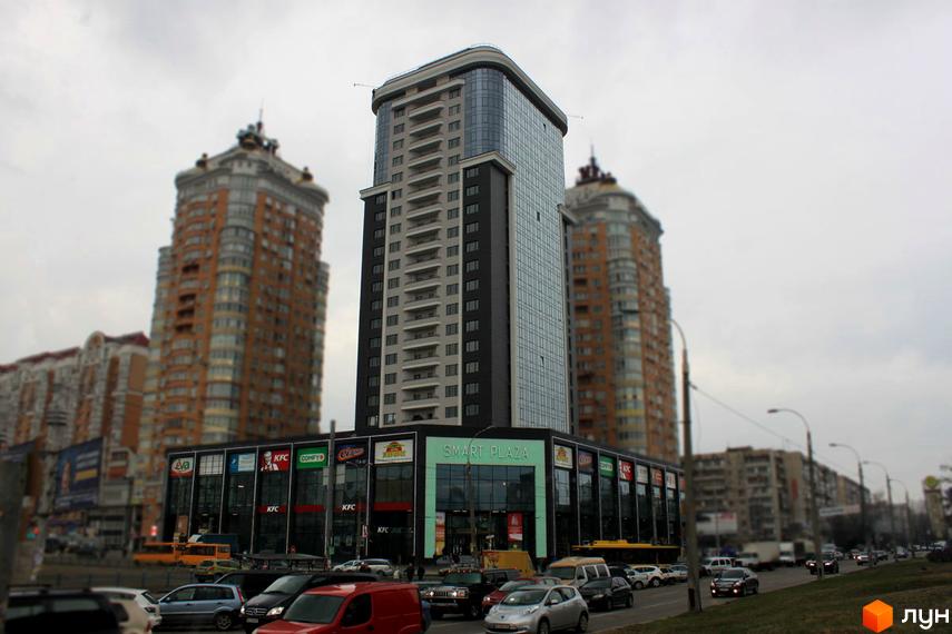 Житловий комплекс Smart Plaza Obolon - це сучасний багатоповерховий будинок з панорамним склінням та комерційними приміщеннями на першому поверсі. Навколо комплексу розташовані дороги та інші будівлі.