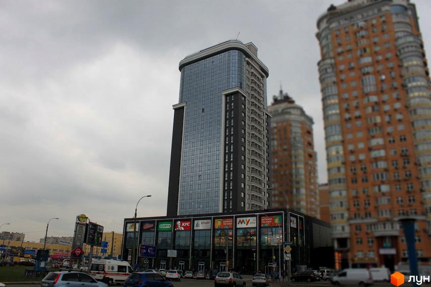 Житловий комплекс Smart Plaza Obolon – висотна будівля з панорамним склінням та комерційними приміщеннями на першому поверсі. Навколо комплексу розташовані дороги та інші будівлі.