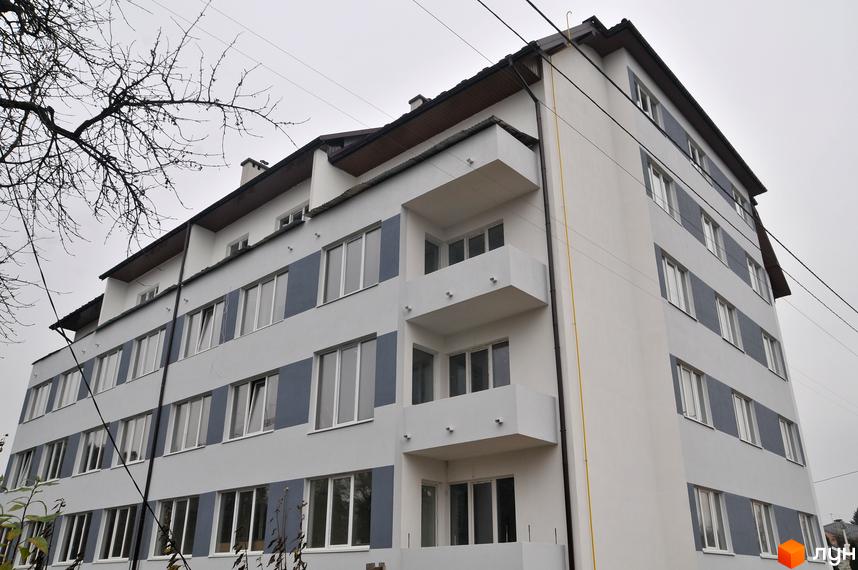 Багатоповерховий будинок з білими стінами та сірими вставками. На фасаді є балкони та великі вікна. На передньому плані видно гілки дерев.