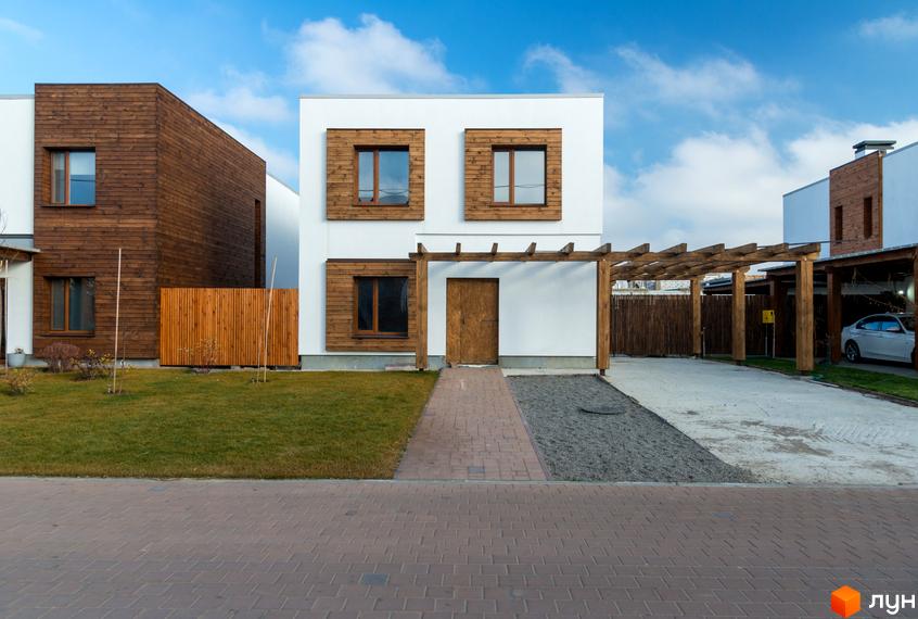 Котеджне містечко КМ На Щасливій: двоповерховий будинок з білою фасадною частиною, дерев'яними вставками та навісом. Прибудинкова територія з газоном, доріжкою та навісом для авто.