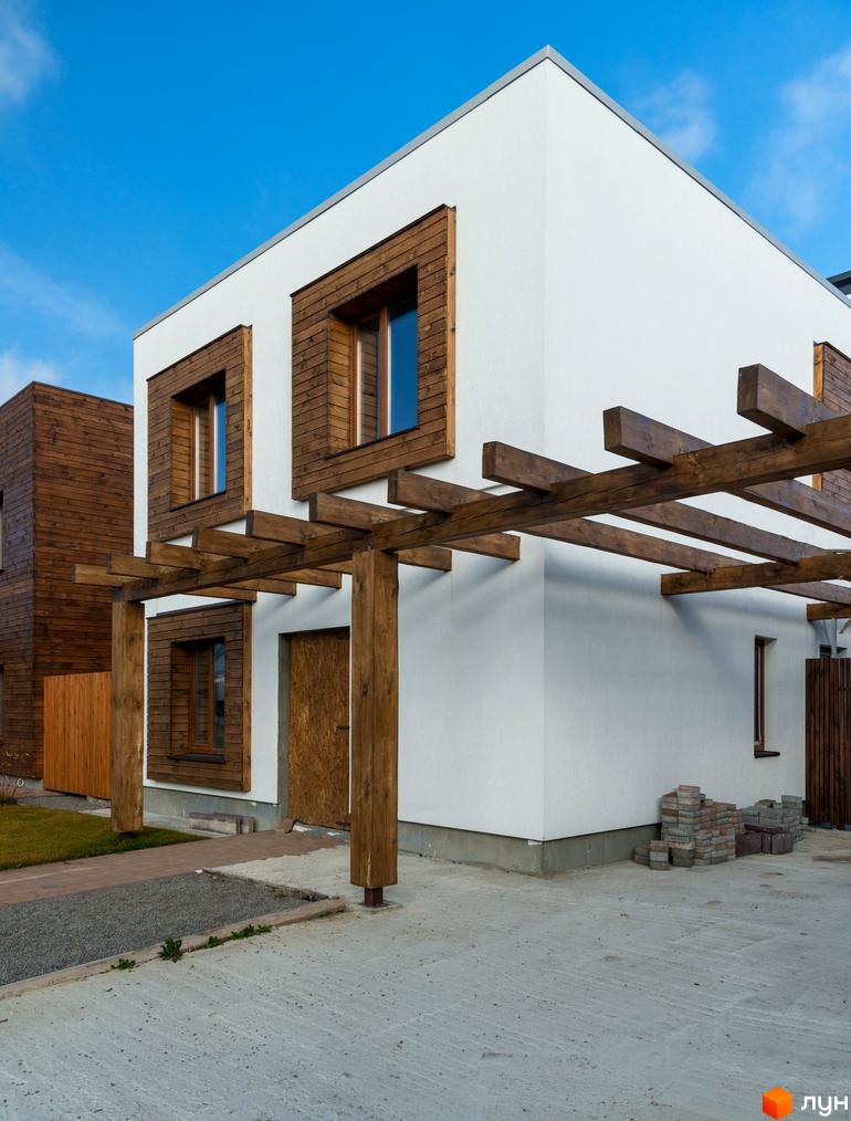 Котеджне містечко КМ На Щасливій. Фасад будинку з білою штукатуркою, дерев'яними елементами та перголою. Поруч облаштований двір з бетонним покриттям та зеленою зоною.