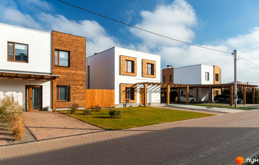 Котеджне містечко На Щасливій: білі будинки з дерев'яними елементами та навісами для авто. Прибудинкова територія з газонами та бруківкою.