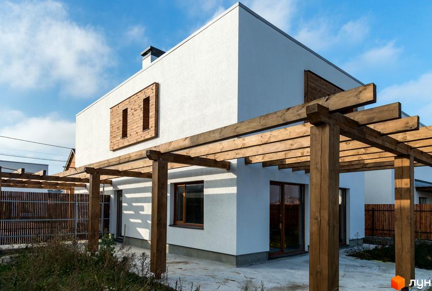 Котеджне містечко КМ На Щасливій. Білий двоповерховий будинок з дерев'яною обшивкою та перголою. Навколо будинку облаштована прибудинкова територія з парканом.