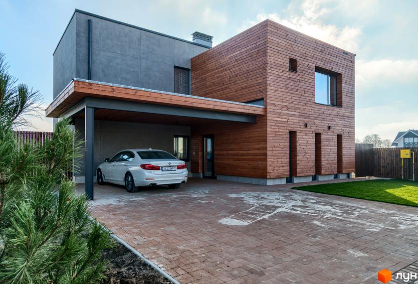 Котеджне містечко На Щасливій: двоповерховий будинок з дерев'яним та сірим фасадом, навіс для авто та вимощена цеглою доріжка. Зелена галявина та паркан на прибудинковій території.