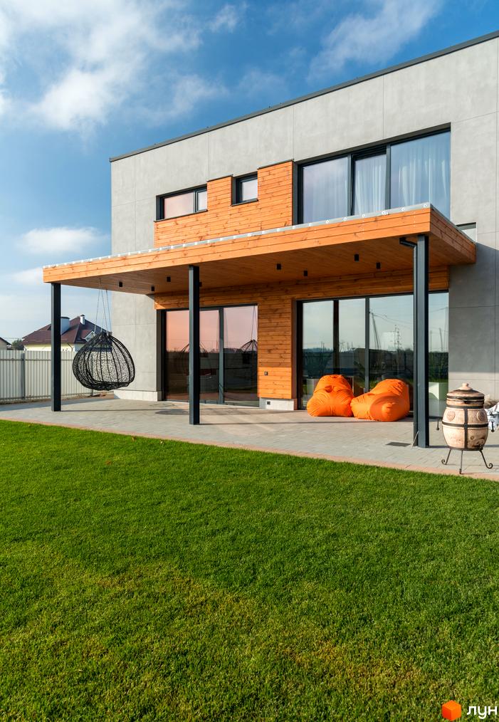 Котеджне містечко На Щасливій: сучасний двоповерховий будинок з дерев'яною терасою та зручним зонуванням. Зелений газон та зона відпочинку з кріслом-гойдалкою та пуфами.