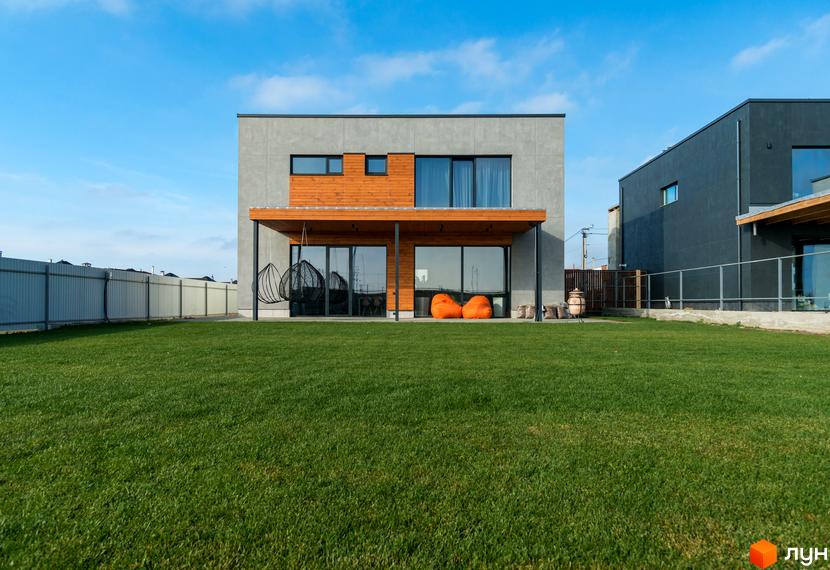 Котеджне містечко На Щасливій. Фасад будинку з сірого бетону та дерев'яними елементами, великі вікна, тераса з підвісними кріслами. Зелений газон перед будинком.