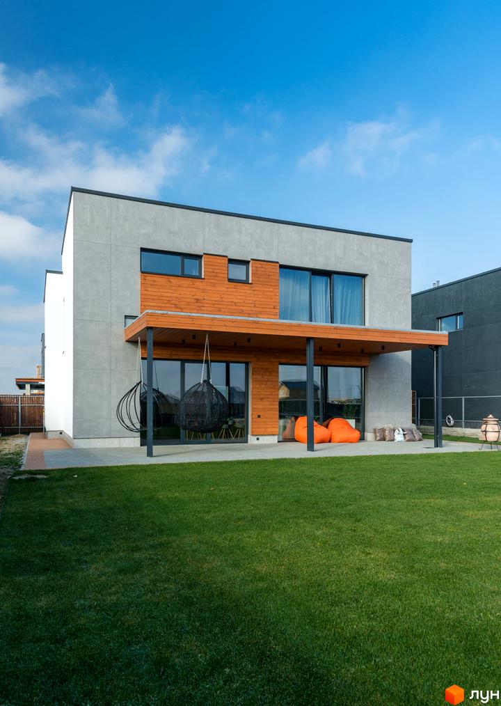 Котеджне містечко КМ На Щасливій: двоповерховий будинок з сірою та дерев'яною обробкою, великими вікнами та навісом. Поруч зелена галявина з кріслами-мішками та гойдалками.