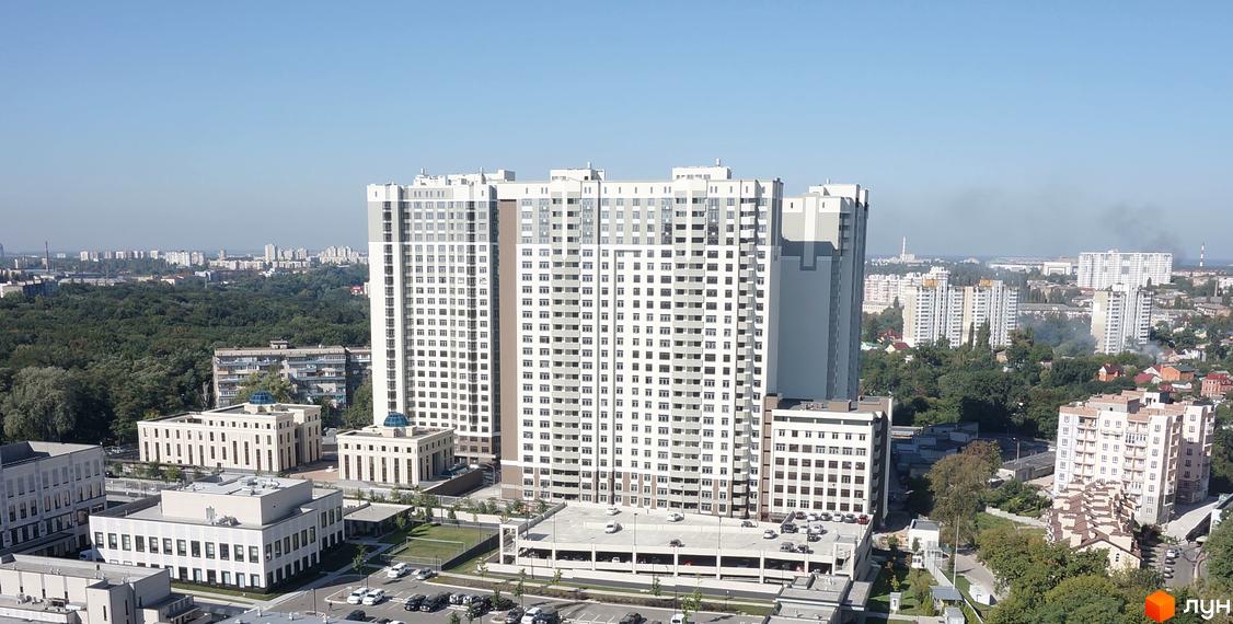 Зелений острів-2 - новий житловий комплекс з високими білими будинками та сірими елементами. Навколо зелені насадження та паркінг з автомобілями.