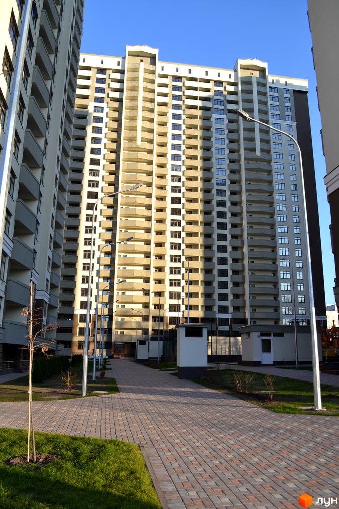 Новобудова Зелений острів-2 з численними балконами та вікнами на тлі блакитного неба. Прибудинкова територія з бруківкою та зеленими насадженнями.