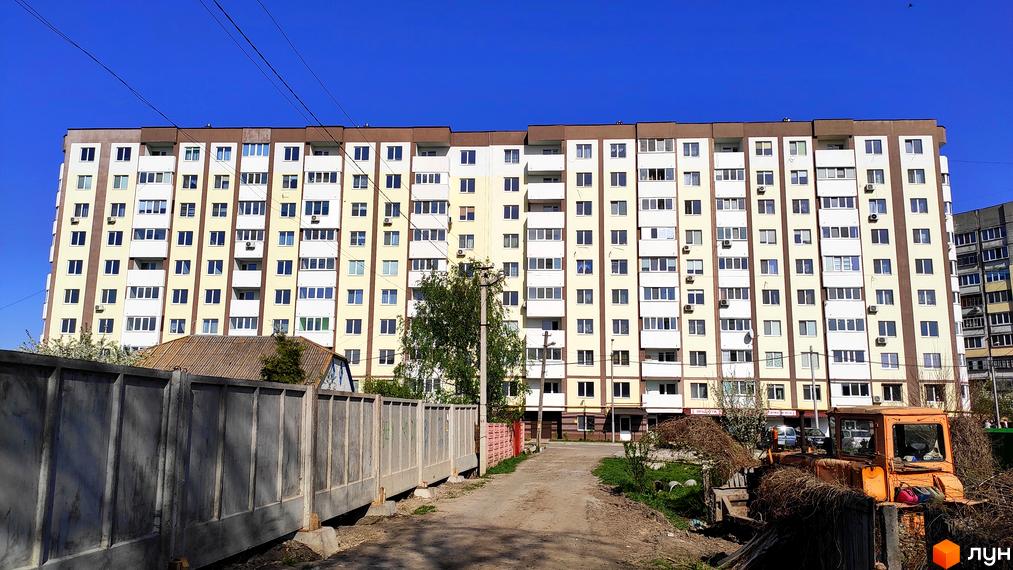 Житловий комплекс Банківський: багатоповерховий будинок світло-жовтого кольору з коричневими вставками. На передньому плані ґрунтова дорога, паркан та стара техніка.