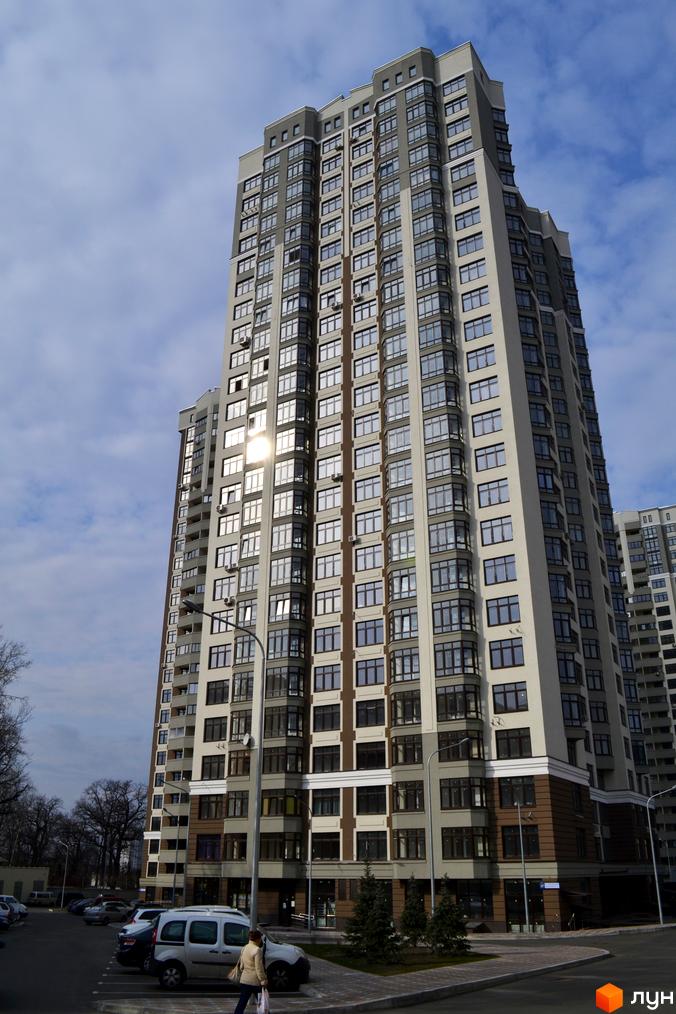 Високоповерховий житловий комплекс Зелений острів-2 з сіро-бежевим фасадом та великою кількістю вікон. Навколо будинку облаштований паркінг та зелені насадження.