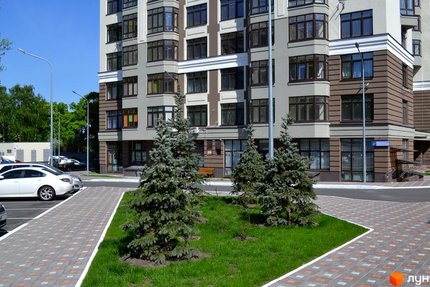 Зовнішній вигляд новобудови Зелений острів-2 з фасадом у світлих тонах та коричневими акцентами. Прибудинкова територія з доглянутим газоном, молодими ялинами та вимощеною плиткою доріжкою.
