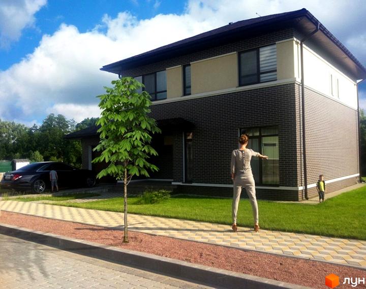 Котеджне містечко КМ Green Town. Двоповерховий будинок з темної цегли та світлої штукатурки. Поряд зелений газон, доріжка з плитки та дерево.