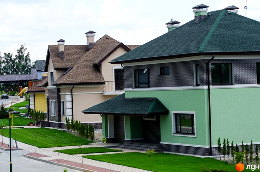 Котеджне містечко КМ Green Town: фасади будинків з зеленим та бежевим оздобленням, сірою цегляною кладкою, коричневими дахами. Доглянута прибудинкова територія з акуратними газонами та доріжками.