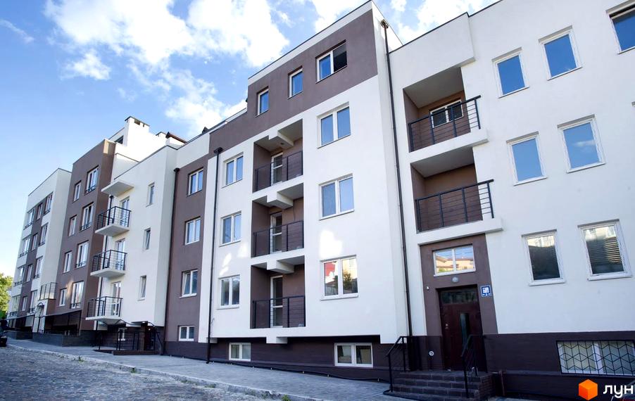 Зовнішній вигляд новобудови ЖК Панорама з білими та коричневими фасадами, балконами та вікнами. Навколо будинку бруківка та невелика прибудинкова територія.