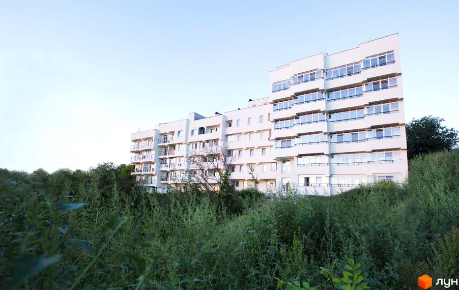 Зовнішній вигляд новобудови ЖК Панорама з численними балконами та великими вікнами. Навколо будинку зелена територія з високою травою.