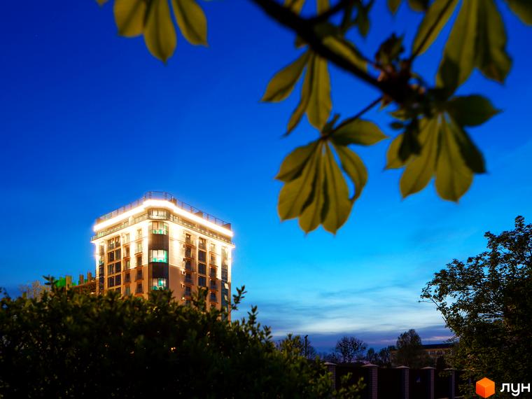 Житловий комплекс HydroPark DeLuxe з підсвіченим фасадом на фоні вечірнього неба. На передньому плані зелені дерева та кущі.