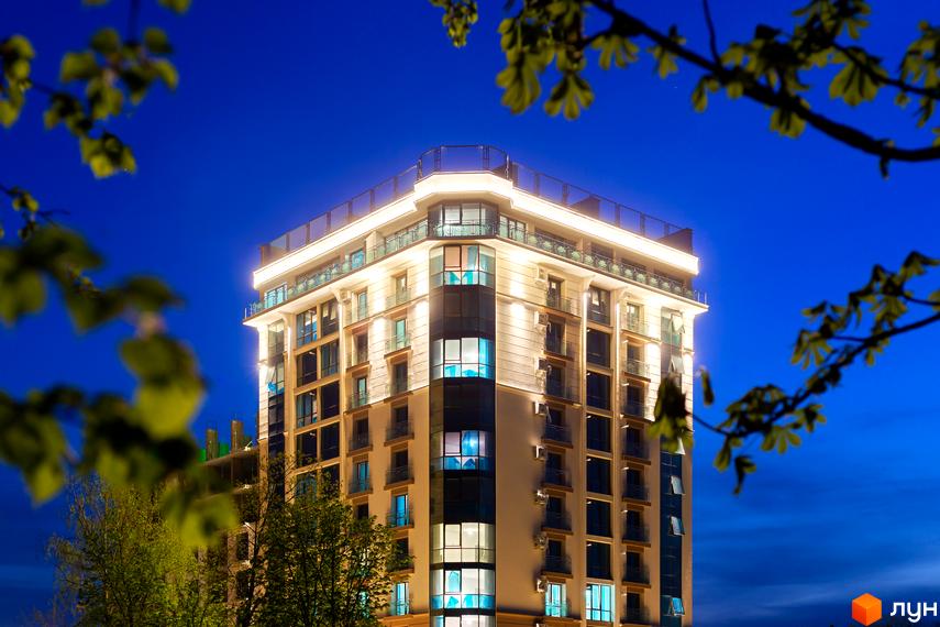Вечірній вигляд житлового комплексу HydroPark DeLuxe з підсвіченим фасадом, вікнами та балконами. Будівля оточена зеленими деревами на тлі синього нічного неба.