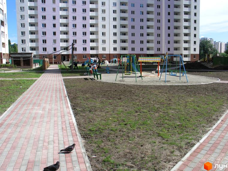 Житловий комплекс на вулиці Урлівській, 38а. Багатоповерховий будинок світло-бузкового кольору з білими балконами. Поруч дитячий майданчик та пішохідна доріжка.