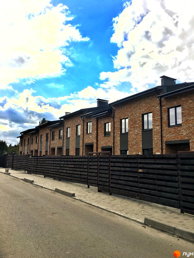 Зовнішній вигляд цегляних таунхаусів „Пушкінський“ з темними дахами та вікнами, що стоять вздовж дерев'яного паркану та бруківки.