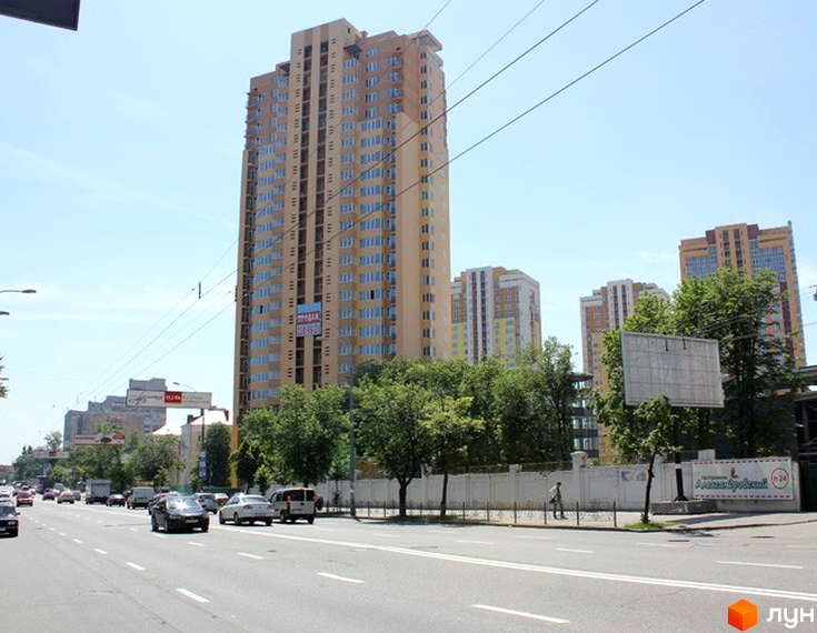 Високий житловий комплекс Червонозоряний світло-коричневого кольору з багатьма вікнами. Поруч широка дорога з автомобілями та зелені дерева.