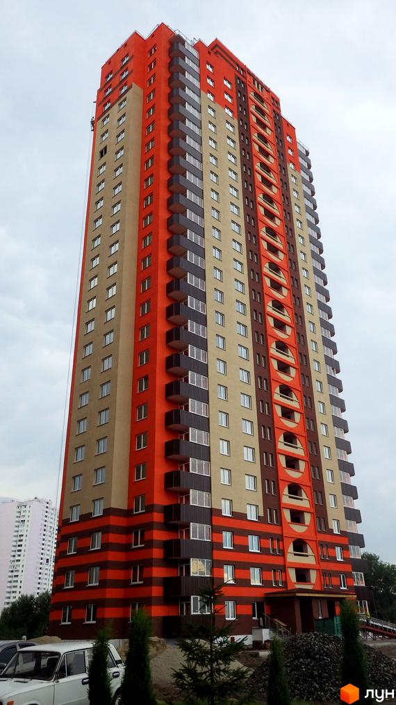 Новобудова на вул. Григоренка, 12а, Позняки-2, має фасад з помаранчевими, бежевими та темно-коричневими секціями, включаючи круглі елементи. Прибудинкова територія містить зелені насадження.