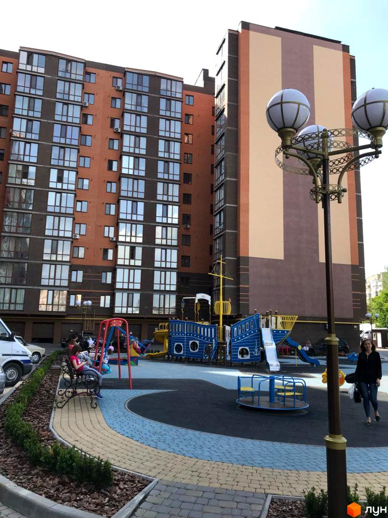Зображення фасаду та дитячого майданчика Житлового комплексу Сімейний. Багатоповерховий будинок з вікнами та ігровий простір для дітей.