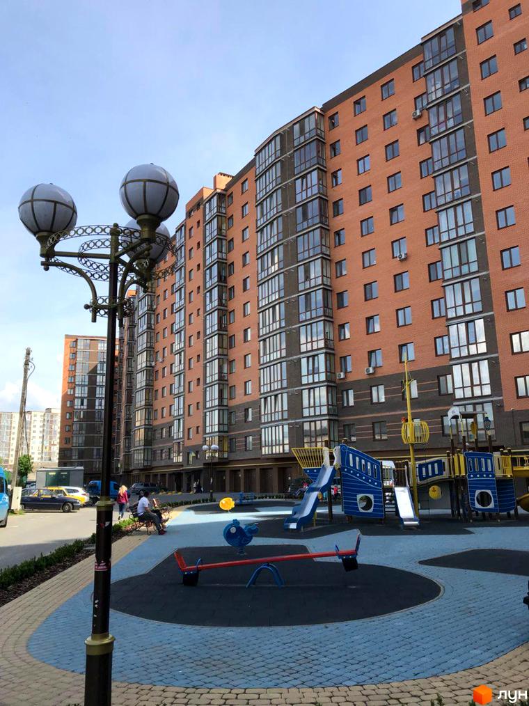 Зображення фасаду та дитячого майданчика Житлового комплексу Сімейний. Багатоповерховий будинок з червоної цегли з великими вікнами.