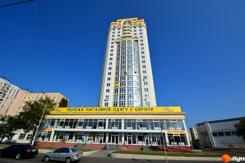 Зображення житлового комплексу Оболонь Тауер. Висотна будівля з магазинами на першому поверсі та автомобілями на вулиці.