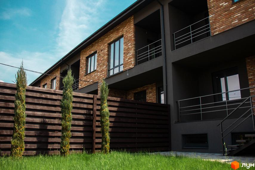 Таунхауси Пушкінський. Зовнішній вигляд цегляних та сірих фасадів з балконами та вікнами. Прибудинкова територія з дерев'яним парканом та зеленим газоном.