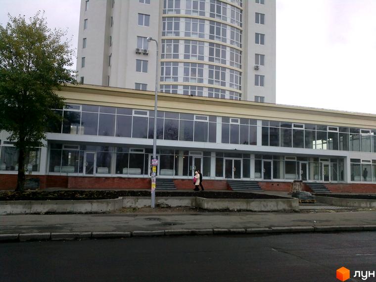 Зображення фасаду Новобудови Оболонь Тауер. Високий будинок з панорамними вікнами та прилеглою територією.