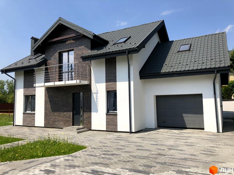 Котеджне містечко КМ Європа: двоповерховий будинок з білою та коричневою цегляною обробкою, балконом та гаражем на тлі блакитного неба. Прибудинкова територія з вимощеним двориком та зеленою травою.