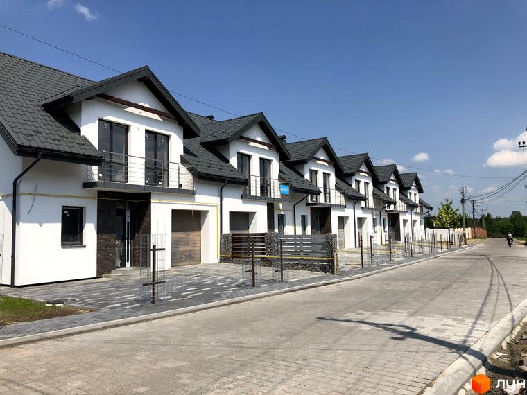 Котеджне містечко КМ Європа: ряд білих двоповерхових будинків з сірими дахами та індивідуальними гаражами. Прибудинкова територія вимощена плиткою, огороджена парканом.