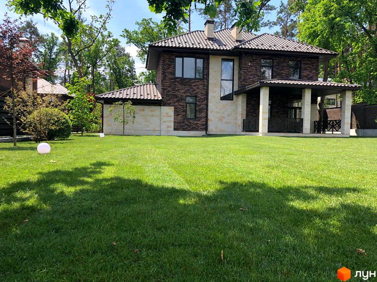 Котеджне містечко Zahrava Village. Двоповерховий будинок з коричневої цегли та світлого каменю з двосхилим дахом. Доглянута зелена галявина перед будинком.