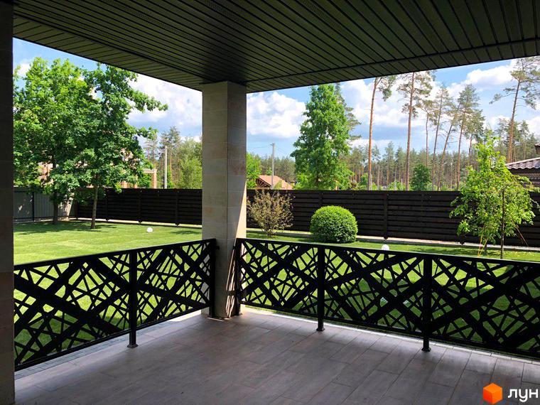 Вид з тераси котеджного містечка КМ Zahrava Village на зелений газон та дерева. Огорожа з чорним ажурним візерунком.