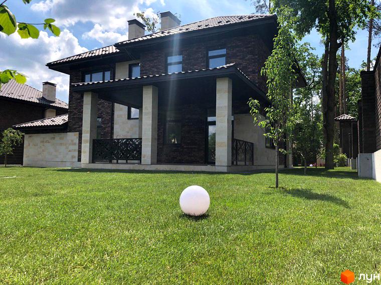 Котеджне містечко Zahrava Village: двоповерховий будинок з коричневої цегли та світлого каменю з навісом та колонами. Доглянутий газон з декоративною кулею перед будинком.