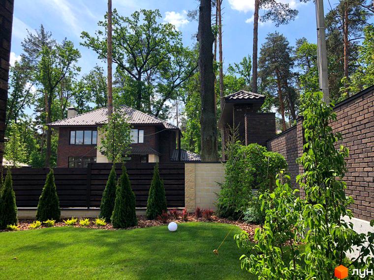 Котеджне містечко Zahrava Village: двоповерховий будинок з цегляним фасадом та великими вікнами, огороджений дерев'яним парканом. Доглянута прибудинкова територія з газоном та туями.