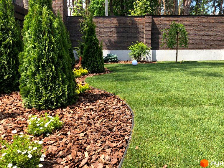 Котеджне містечко Zahrava Village: зелений газон та доглянута прибудинкова територія з туями та декоративними рослинами. Цегляний паркан та дерева на задньому плані.