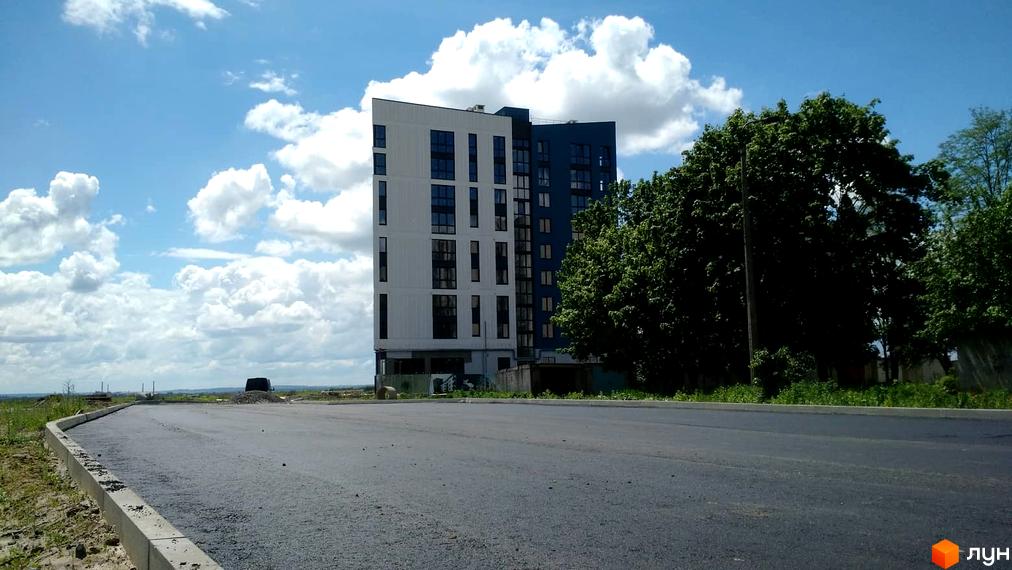 Новобудова ЖК Басів схил з білою та синьою фасадною частиною на фоні хмарного неба. Поруч розташована дорога з бордюром та зелені дерева.