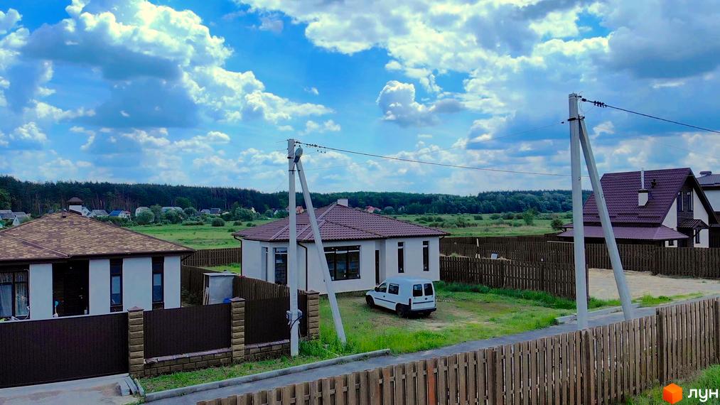Котеджне містечко Михайлівський хутір. Сучасні будинки з коричневими дахами та парканами на тлі зеленого поля та блакитного неба з хмарами. Білий мінівен стоїть біля одного з будинків.