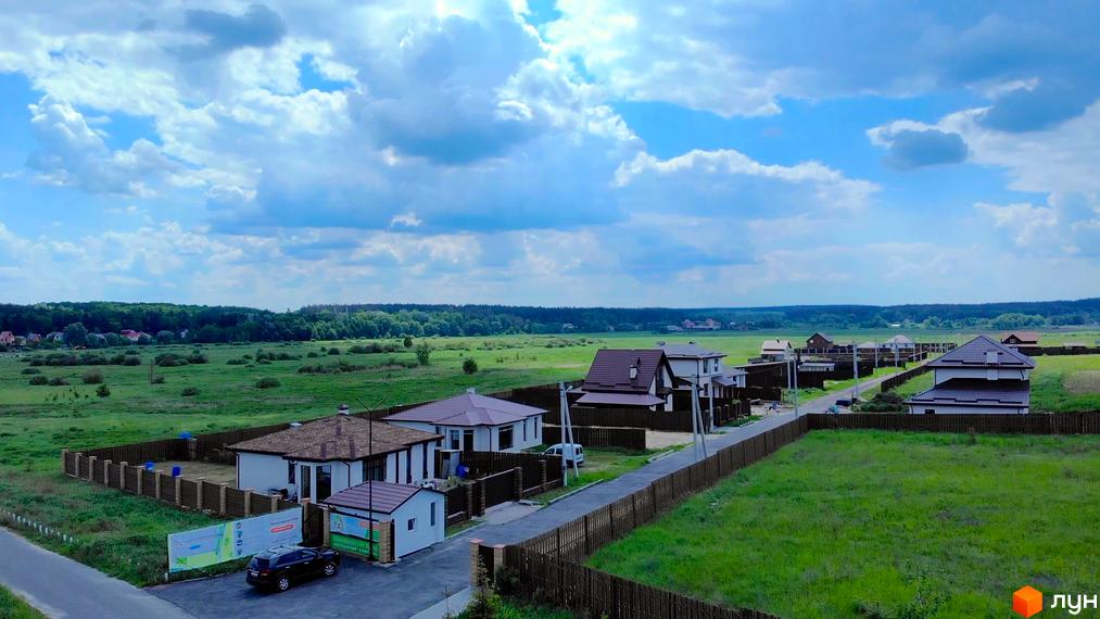 Котеджне містечко Михайлівський хутір: сучасні будинки з коричневими дахами та огородженими ділянками на тлі зеленого поля та лісу. На території комплексу видно дорогу та паркан.